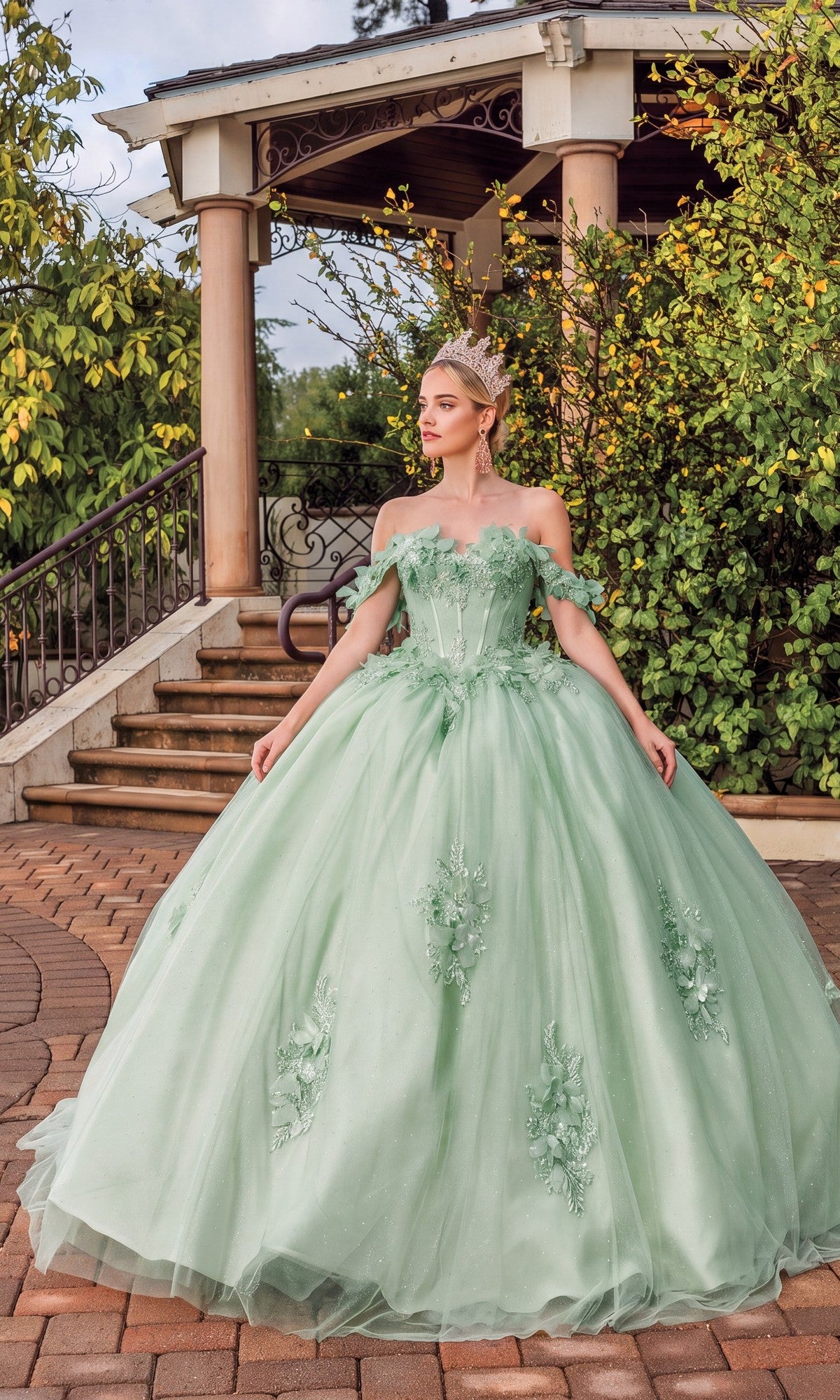A woman in the Quinceanera Dress 1883 By Dancing Queen—a sage ball gown with a sweetheart neckline—wears a sparkling tiara as she stands on a brick path before a gazebo surrounded by greenery.